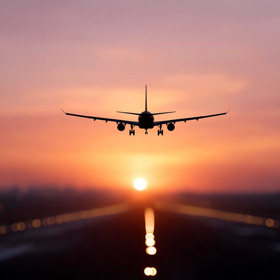 Commercial airplane descending toward a runway at sunset, silhouetted against a pink-orange sky with the sun low on the horizon and runway lights blurred below.