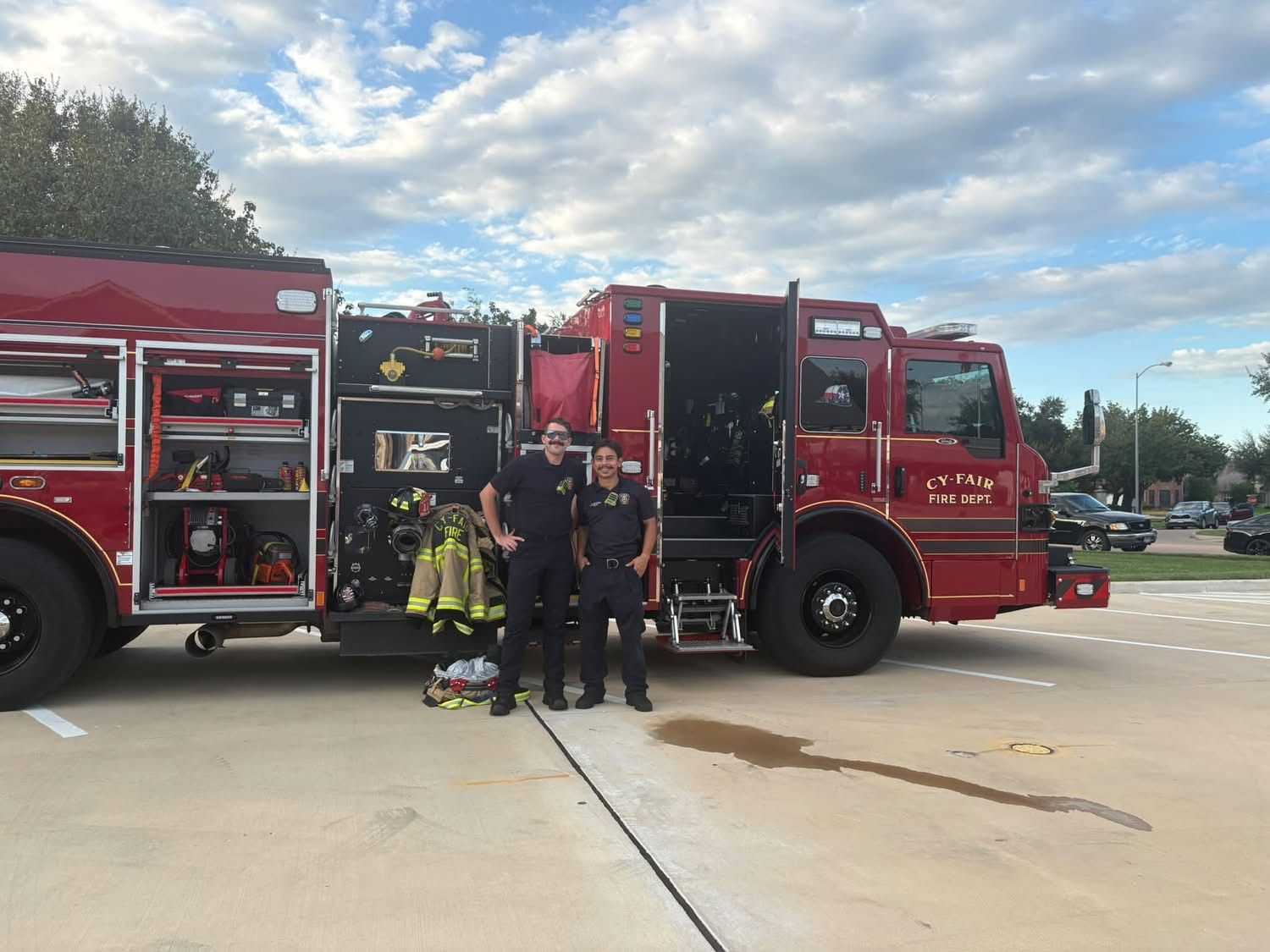 Two firefighters stand beside an open Cy-Fair Fire Department engine, displaying emergency response equipment and protective gear during a community event in a parking lot.