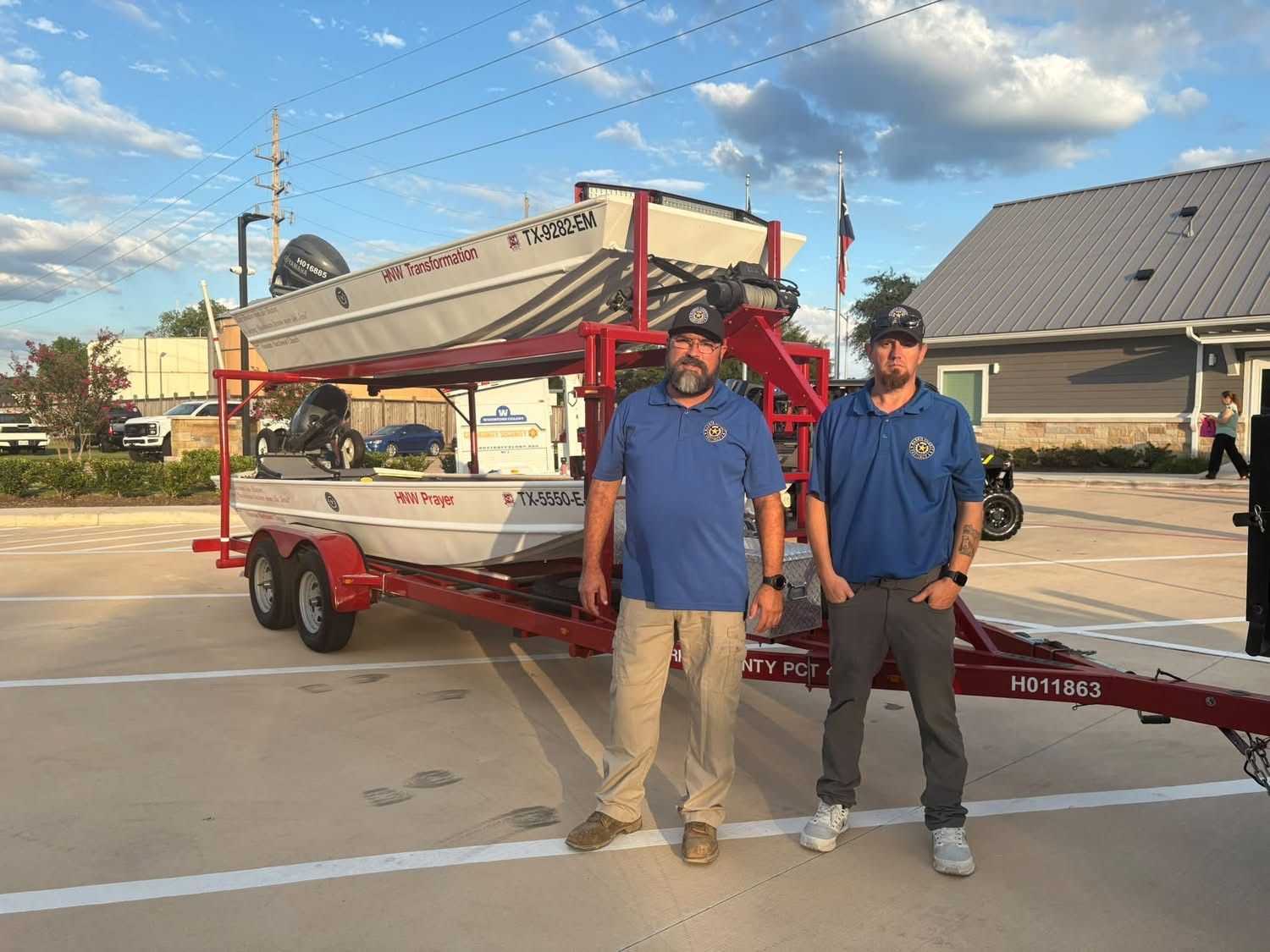 Two emergency response personnel stand beside a trailer carrying rescue boats in a parking lot, displaying water rescue equipment during a community safety event.