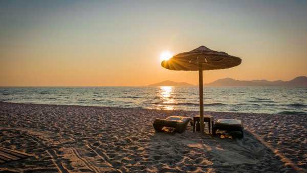 Two empty lounge chairs beneath a thatched beach umbrella on sandy shore at sunset, with calm ocean water, sun reflections, and distant mountains along the horizon.