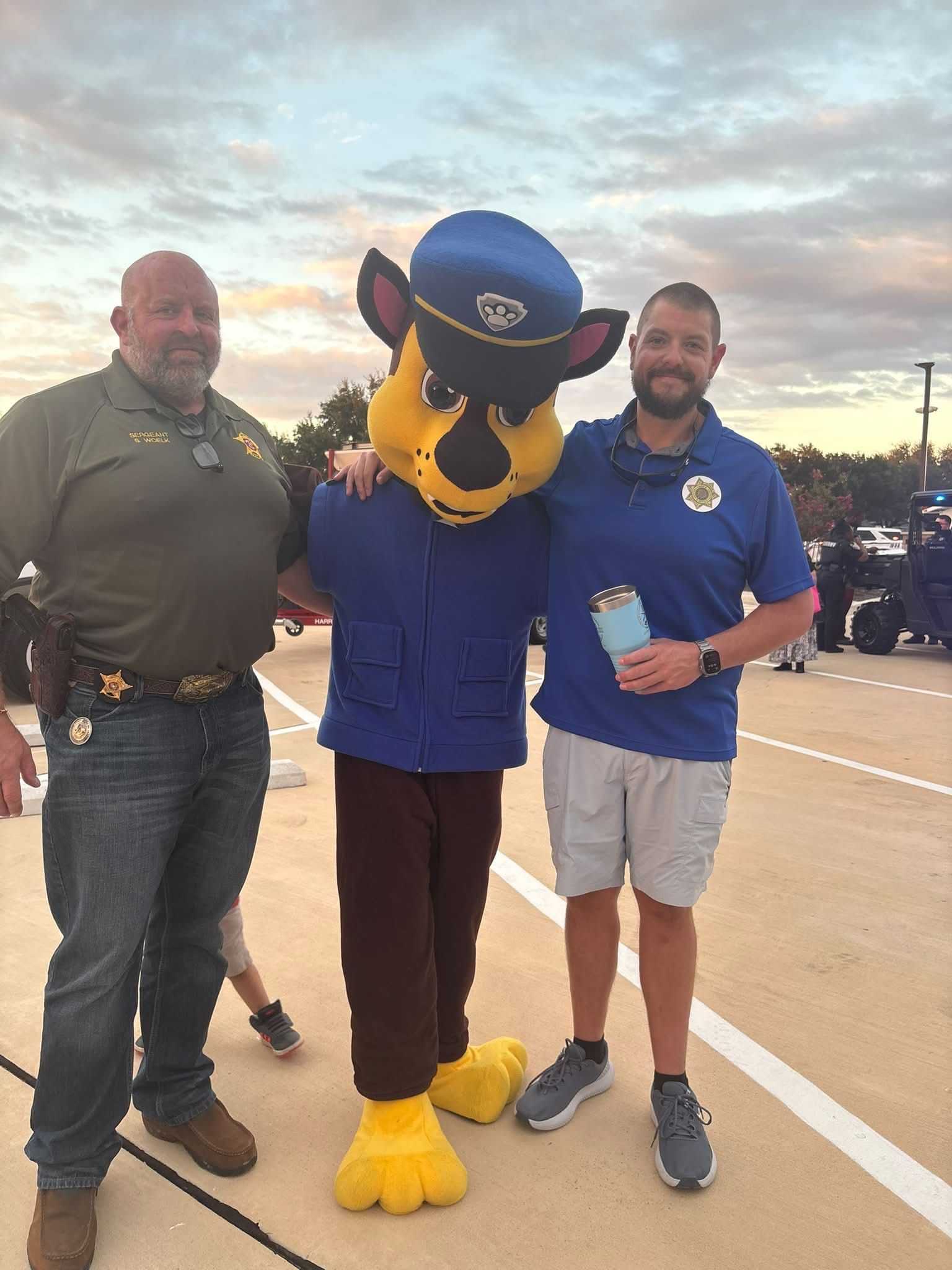 Two community members pose with a police mascot character in a parking lot during a public safety event, with emergency vehicles and attendees visible in the background.