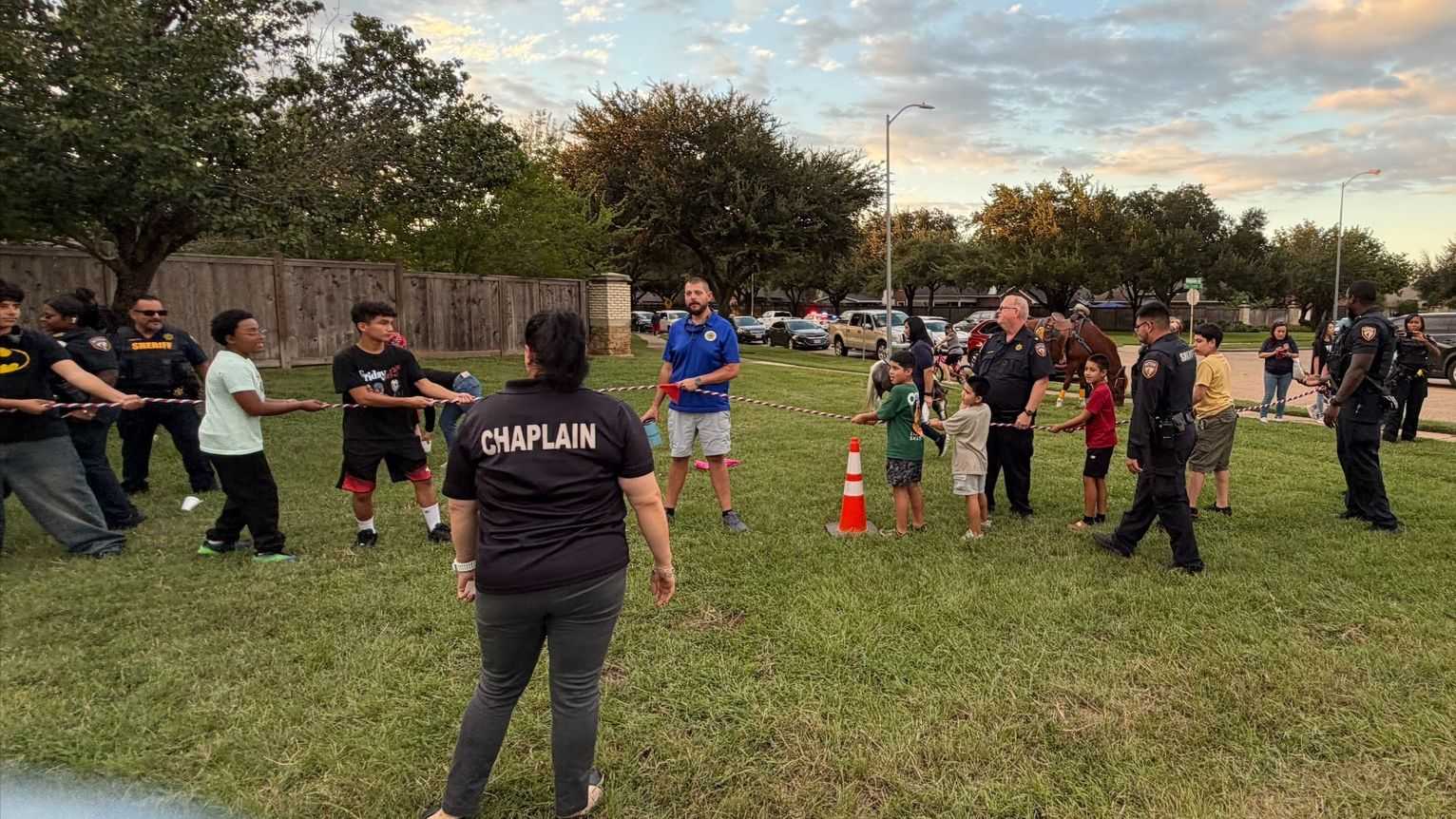 Community event on a grassy field where children, sheriff’s deputies, and volunteers participate in a tug-of-war game, with residents, patrol vehicles, and a mounted deputy nearby.