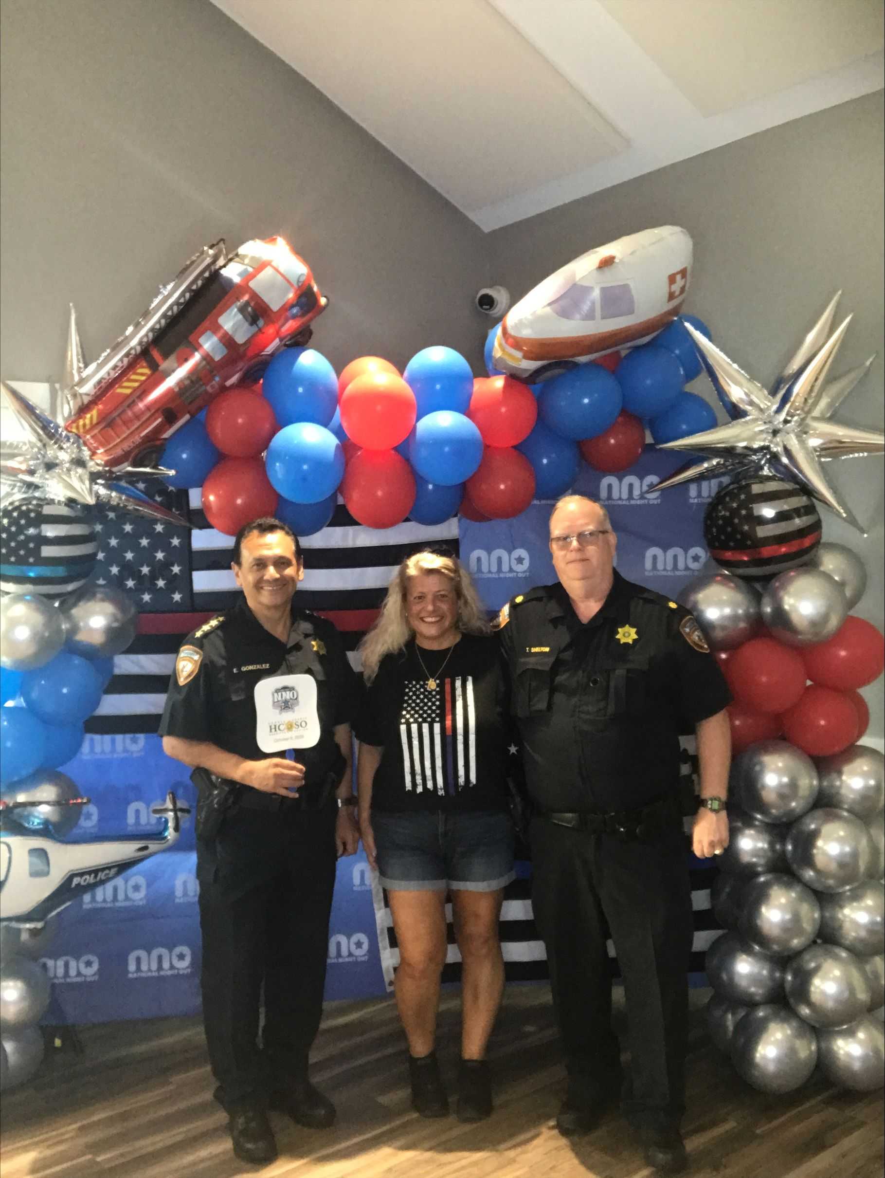 Three attendees pose in front of a National Night Out photo backdrop decorated with red, white, blue, and silver balloons, plus police, fire, and EMS-themed balloon displays.