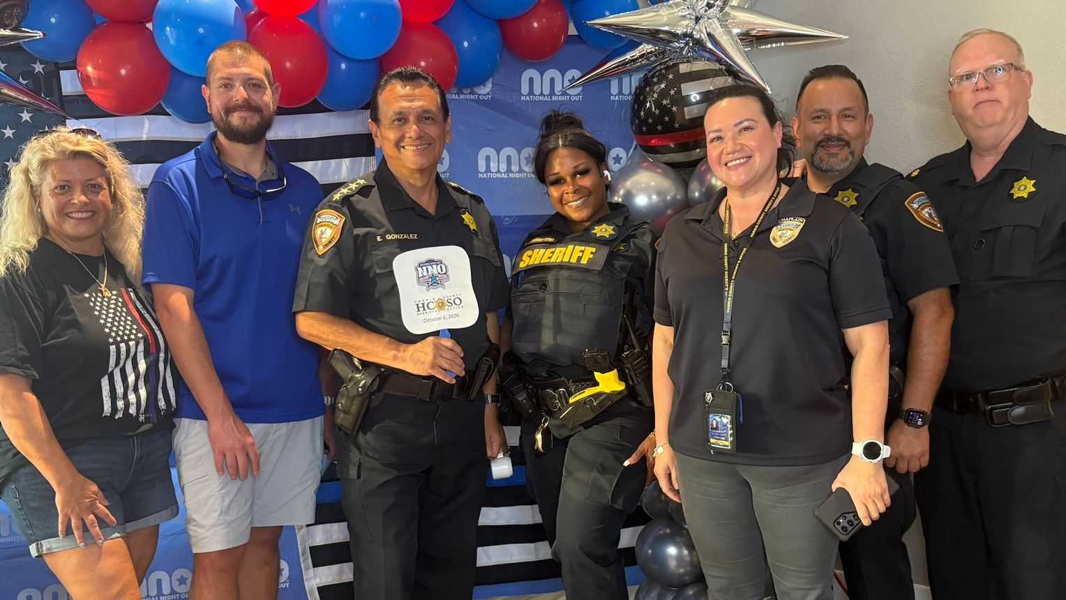 Group photo of community members, sheriff’s deputies, and volunteers posing in front of a National Night Out backdrop decorated with red, white, blue, and silver balloons.