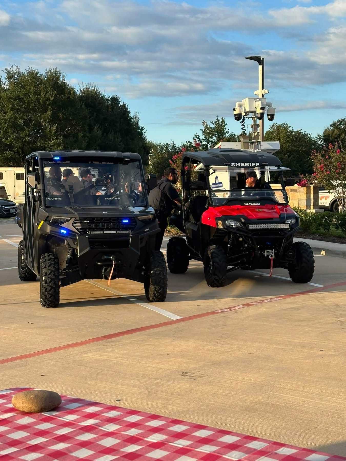 Two sheriff’s utility vehicles with emergency lights are parked at a community safety event, with deputies and surveillance equipment visible in the background.