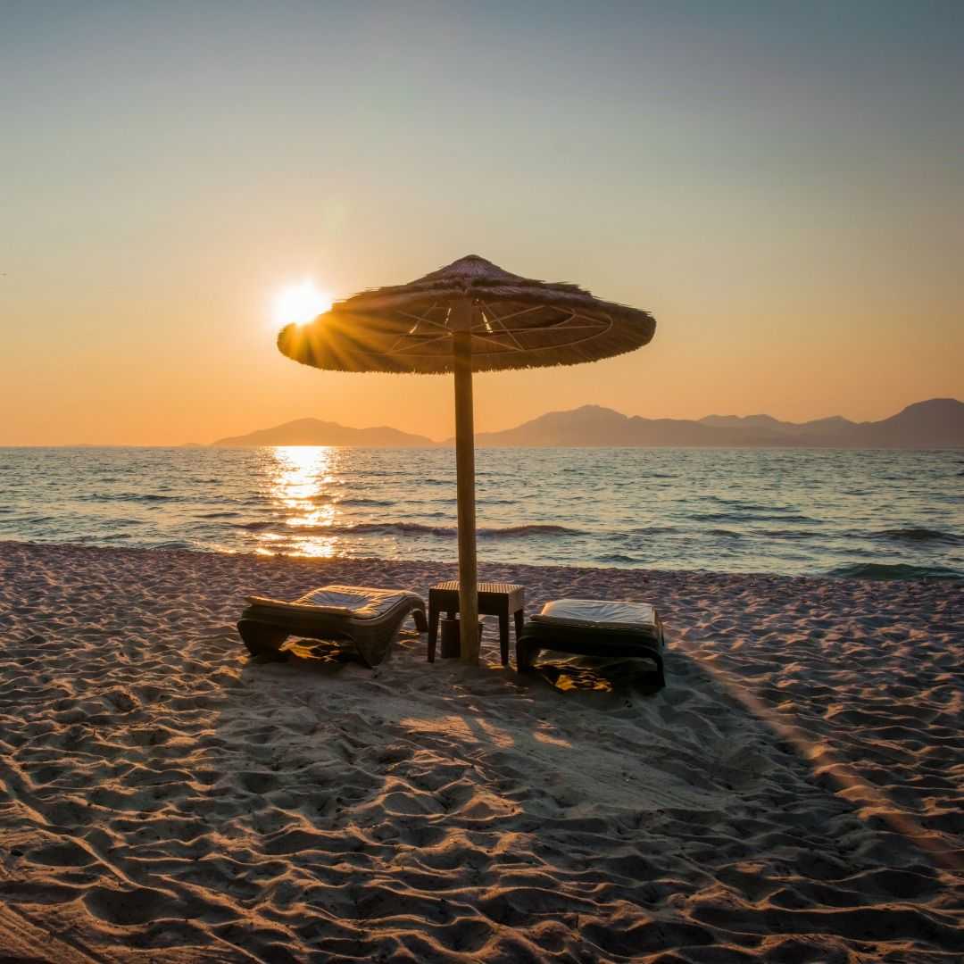 Two empty lounge chairs beneath a thatched beach umbrella on sandy shore at sunset, with calm ocean water, sun reflections, and distant mountains along the horizon.