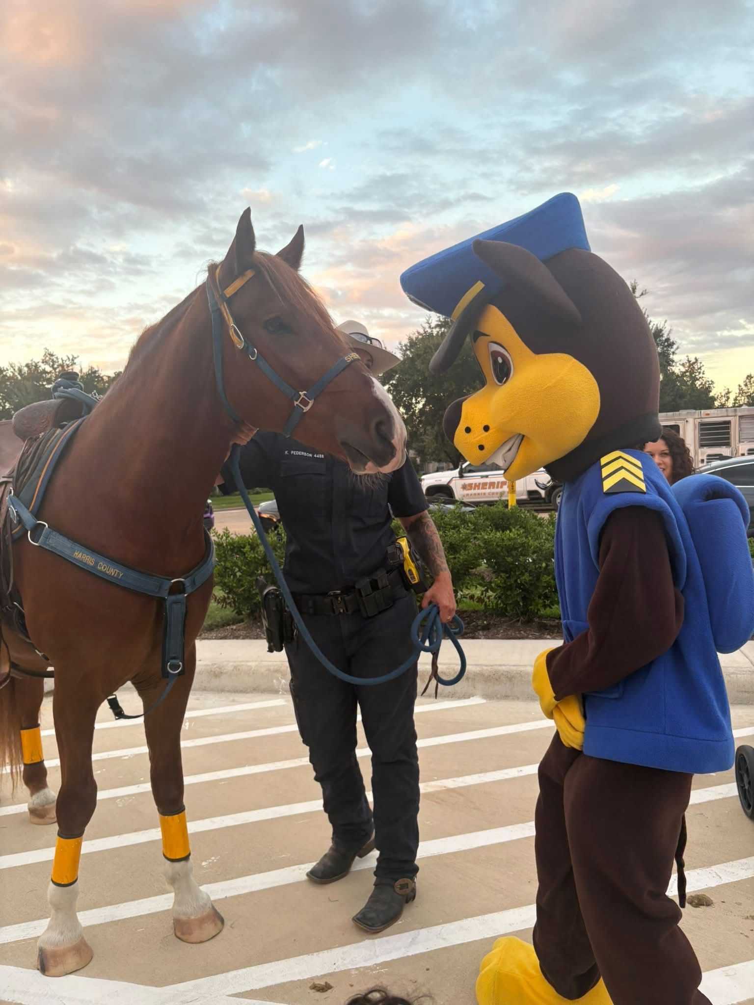 Mounted sheriff’s horse and a police mascot character face each other at a community event, while a deputy holds the horse’s lead in a parking lot at sunset.