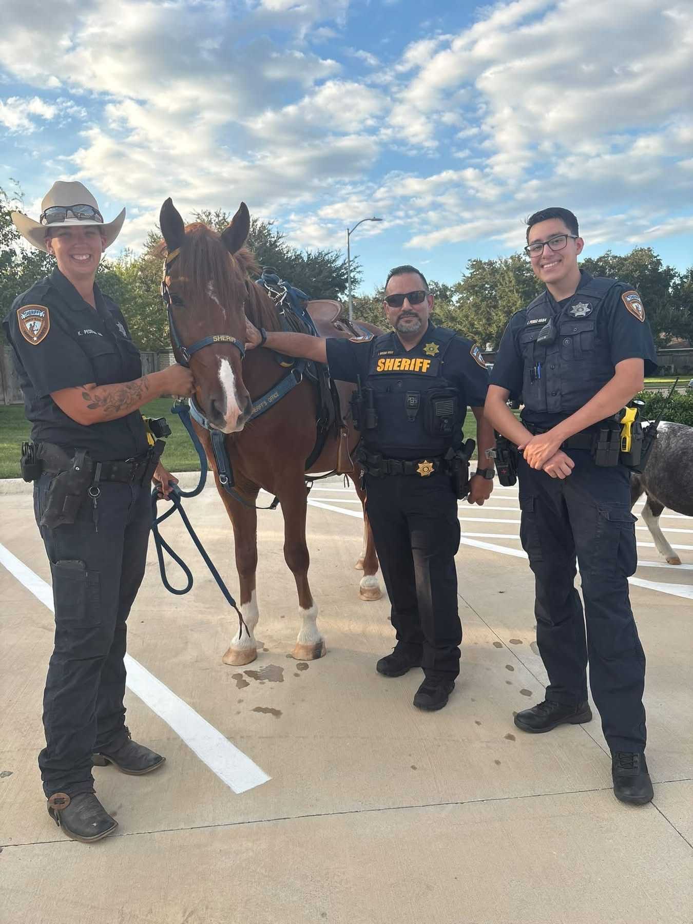 Three sheriff’s deputies stand with a mounted patrol horse in a parking lot during a community event, with the horse wearing a saddle and law enforcement tack.